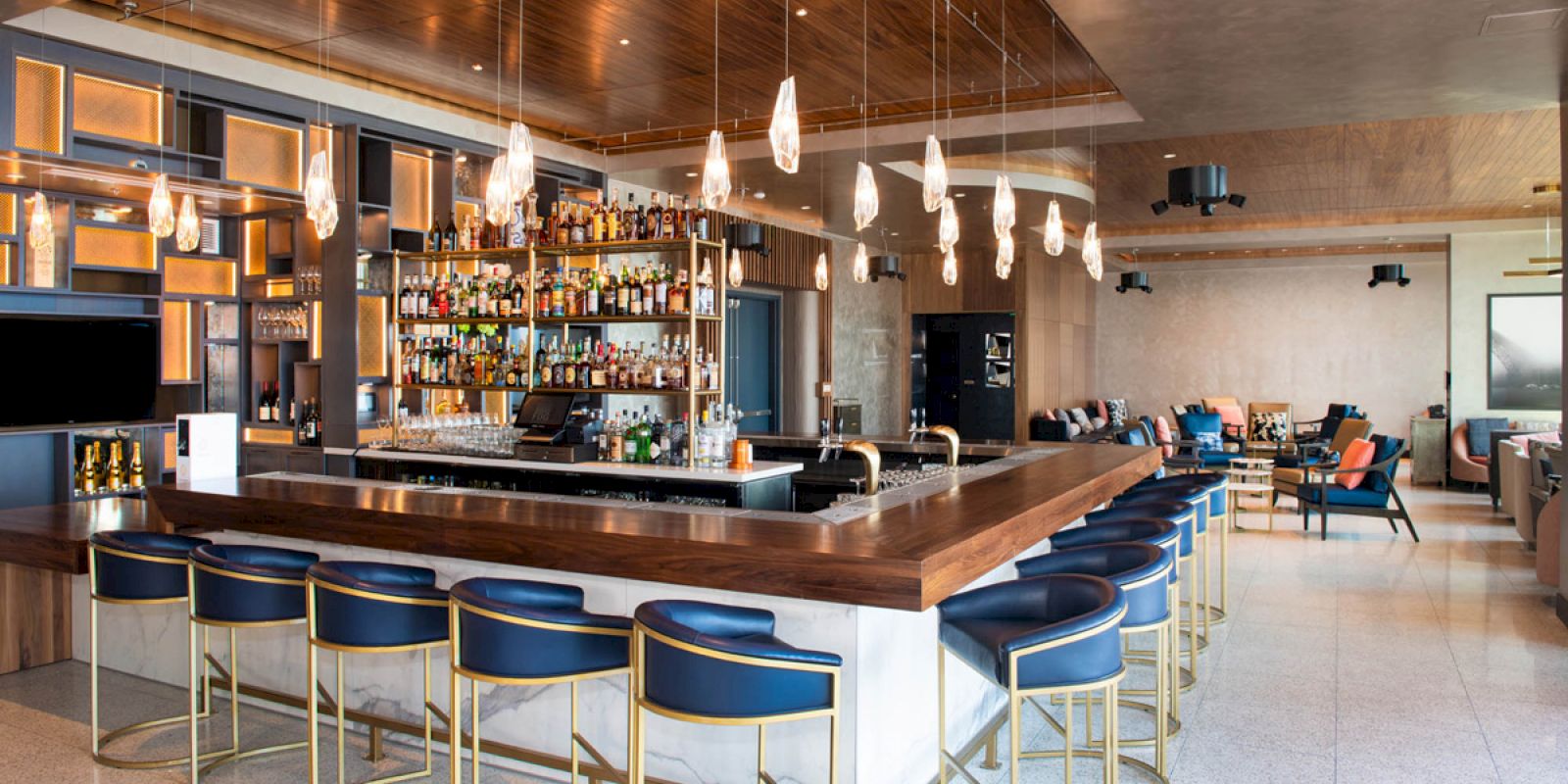 A stylish bar area with a wooden counter, modern lighting, blue bar stools, and a well-stocked shelf of beverages, along with lounge seating.