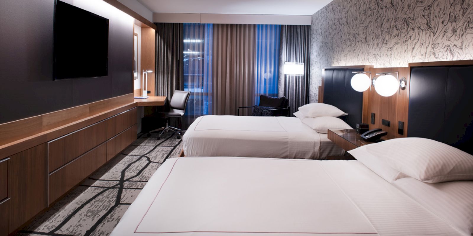 A modern hotel room with two neatly made beds, a wall-mounted TV, a desk with a chair, and stylish lighting fixtures on the wall.
