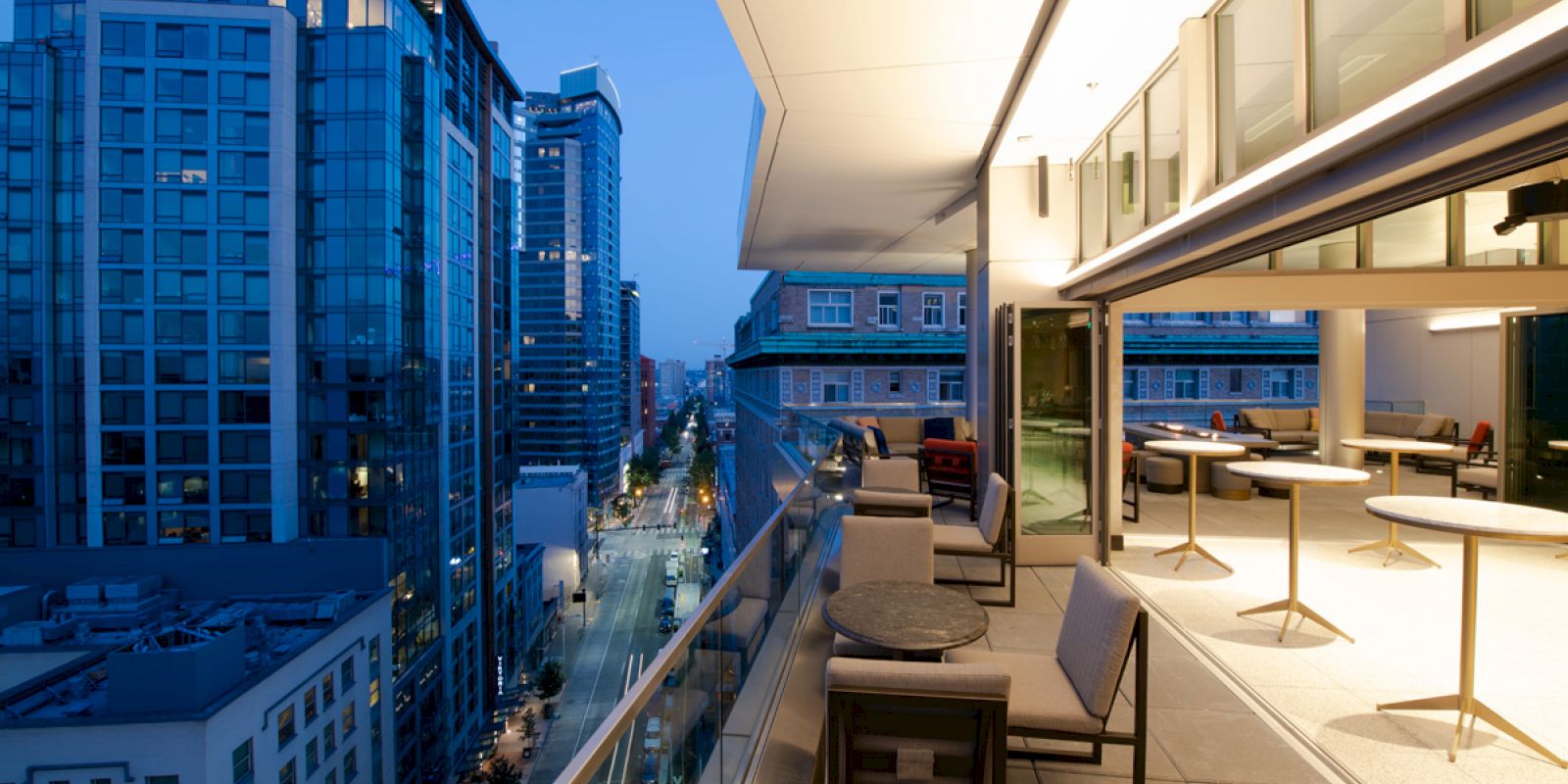 A modern outdoor patio with tables and chairs overlooks a cityscape with tall buildings and a street lit up in the early evening.