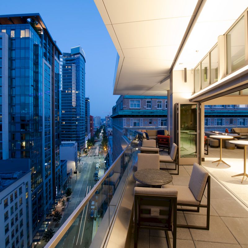 A modern high-rise balcony at dusk, showing a cityscape with tall buildings, outdoor seating, and tables. The area is brightly lit.