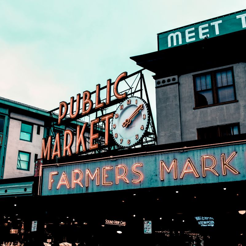 The image shows the exterior of a building with a neon sign that reads "Public Market - Farmers Market" and a clock, suggesting a bustling market location.