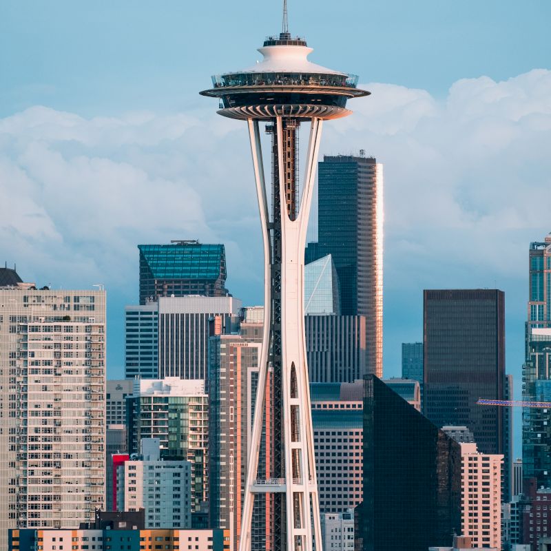The image shows the iconic Space Needle with tall buildings in the background under a partly cloudy sky. It appears to be taken in Seattle.