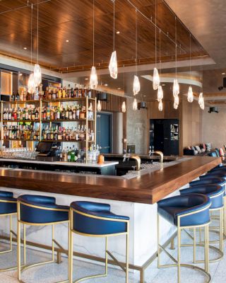The image shows a modern bar with a wooden countertop, stylish hanging lights, and blue bar stools. Shelves with bottles are behind the counter.