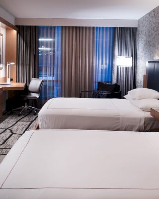 A modern hotel room with two neatly made beds, a flat-screen TV, a workspace, and stylish decor, illuminated by soft lighting.