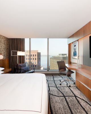 A modern hotel room with a large bed, a wall-mounted TV, a desk with a chair, and a window offering a view of buildings and water.
