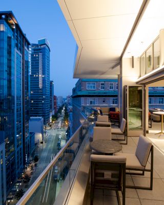 A modern city balcony at twilight with tables, chairs and a stunning view, adjacent to high-rise buildings illuminated by streetlights.
