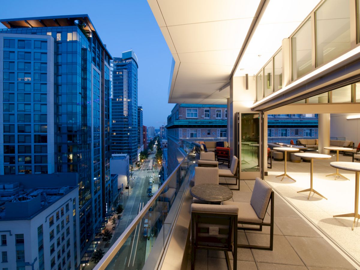 The image shows a modern rooftop balcony at dusk, with seating areas and a view of high-rise buildings and a lit-up street.