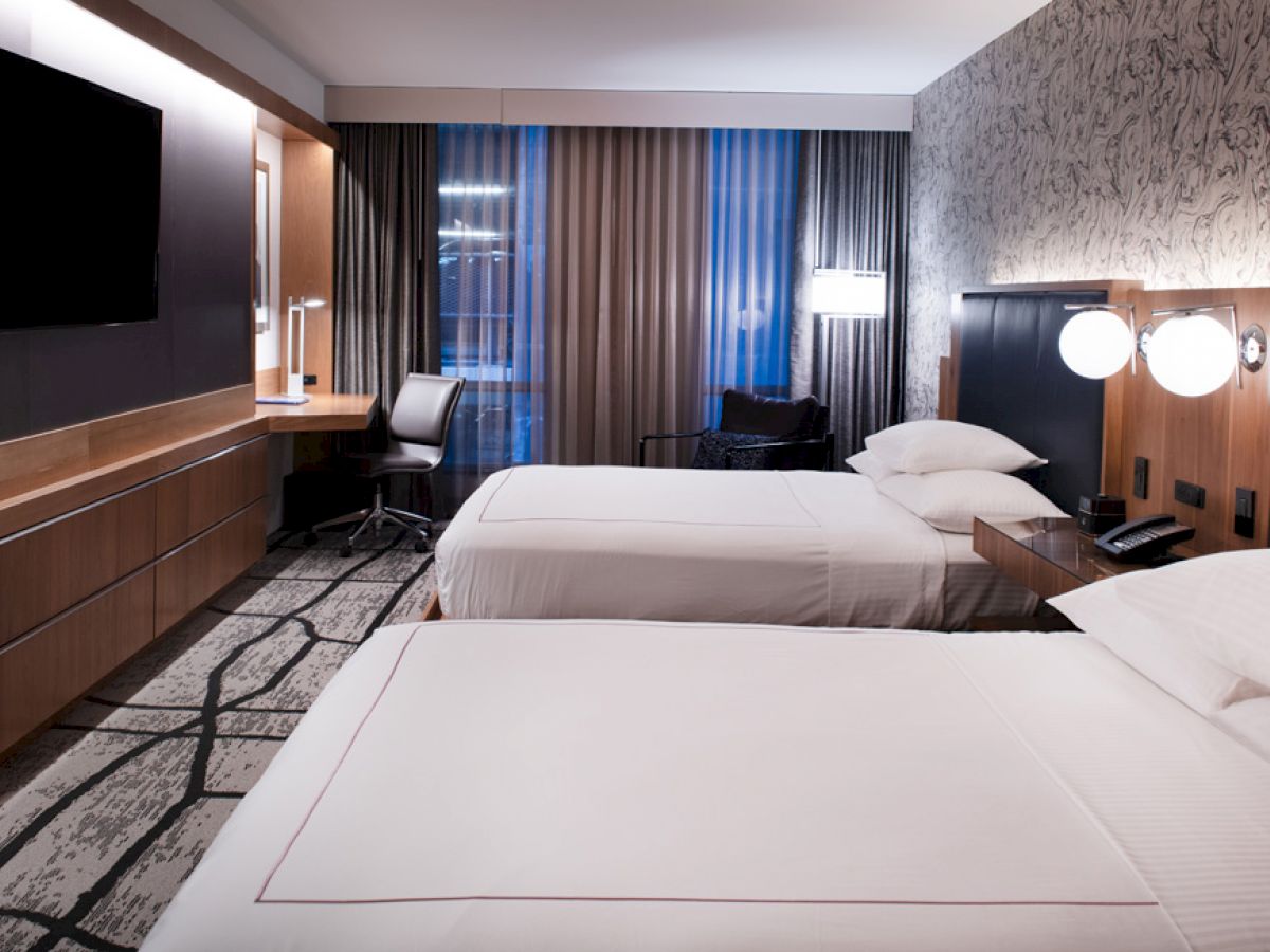 The image shows a modern hotel room with two double beds, a large TV, a desk with a chair, and stylish lighting and decor on walls and floors.