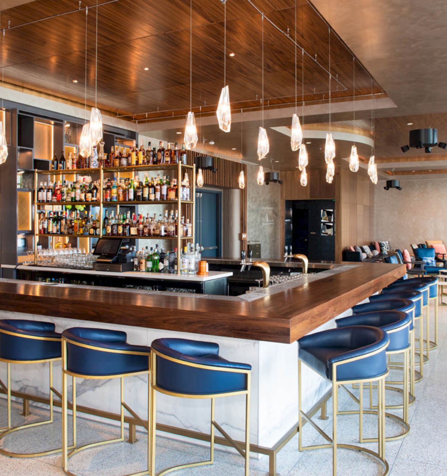 A modern bar with blue and gold stools, hanging lights, and a well-stocked liquor shelf, set in a spacious and stylish interior.