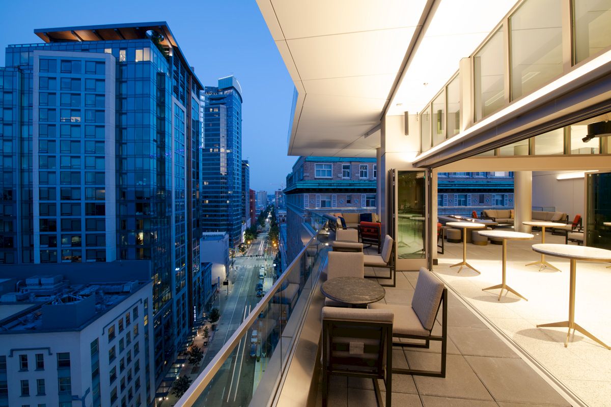 The image shows a modern cityscape at twilight with tall buildings, and a sleek rooftop terrace featuring tables, chairs, and a view of the street below.