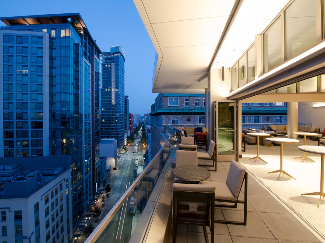 The image shows a modern cityscape at twilight with tall buildings, and a sleek rooftop terrace featuring tables, chairs, and a view of the street below.