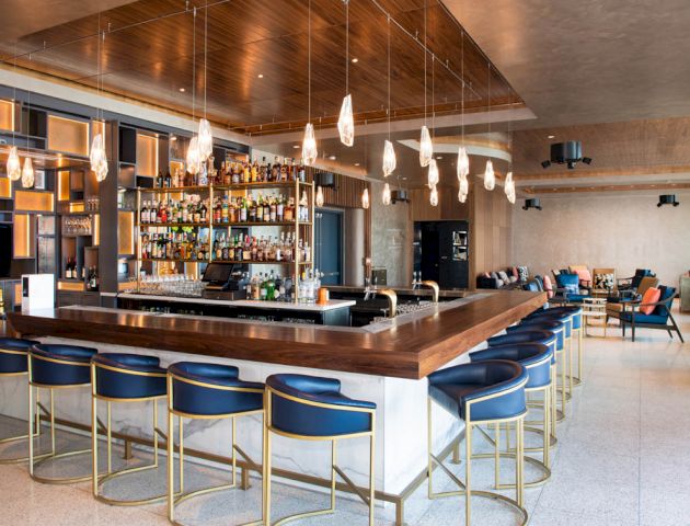 The image shows a modern bar with a large wooden counter, blue cushioned bar stools, various bottles on display, and a stylish, well-lit interior.
