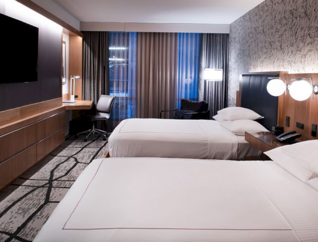 A modern hotel room with two beds, a TV on the wall, a work desk, and a large window with curtains. The room has contemporary decor ending the sentence.