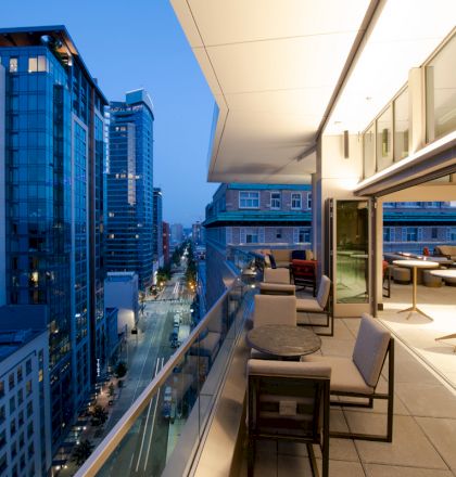 This image shows a modern cityscape at dusk, with tall buildings and a sleek, furnished balcony overlooking the street and skyline.