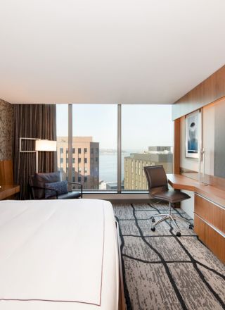 A modern hotel room featuring a large bed, desk, chair, flat-screen TV, and a window view of buildings and water in the background.