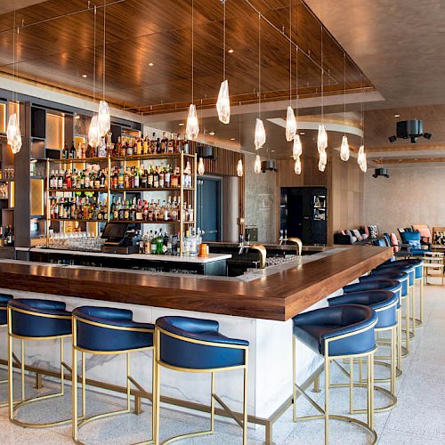 The image depicts a modern bar with a wooden counter, blue cushioned bar stools, hanging lights, and a well-stocked bar area in the background.