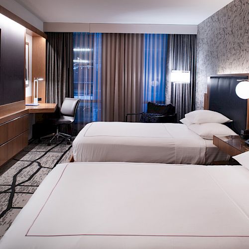 A hotel room with two neatly made beds, a wall-mounted TV, a desk with a chair, and large windows with curtains, creating a cozy atmosphere.