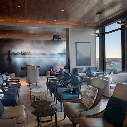 A modern lounge with large windows showcasing a waterfront view, many comfortable chairs, and a cityscape mural on one wall.