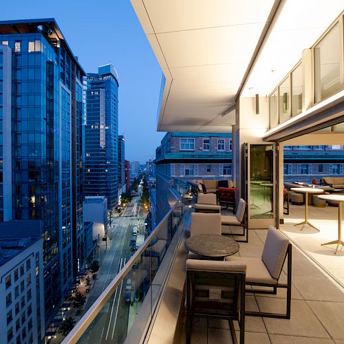 A modern balcony at dusk with outdoor furniture, overlooking city buildings and a street below. Indoors are tables and chairs under warm lighting.