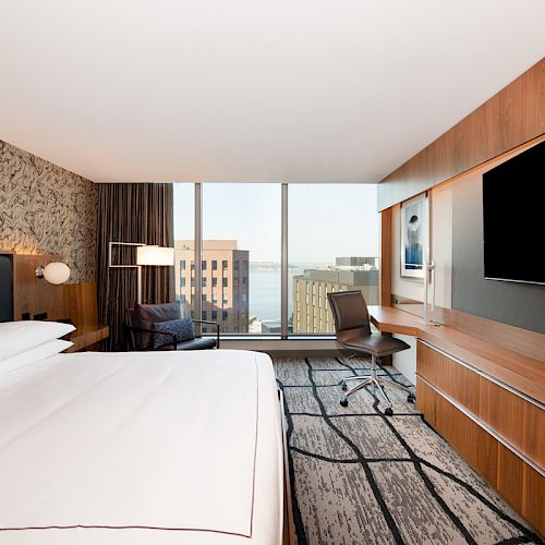 A modern hotel room features a large bed, a wall-mounted TV, a desk, and a view of city buildings through a large window.