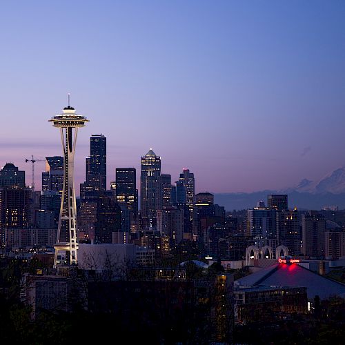 The image shows the Seattle skyline at dusk with the Space Needle prominently visible. Mount Rainier is visible in the background.