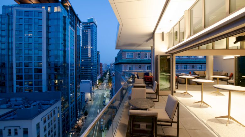 The image shows a modern cityscape with tall buildings at dusk, viewed from a balcony with outdoor seating and tables.