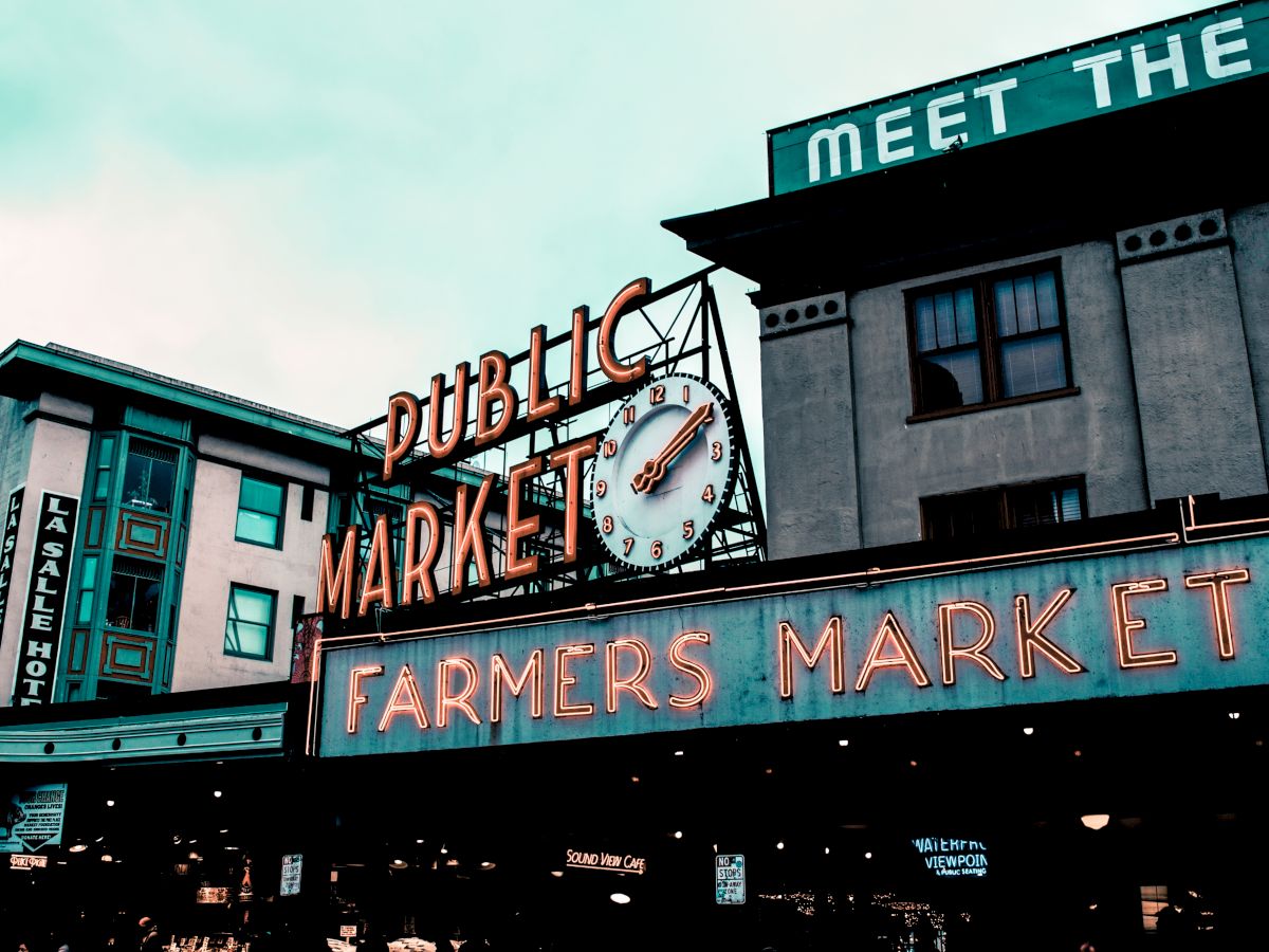 The image shows an illuminated sign reading "Public Market" and "Farmers Market" on a building facade with a clock above it, presumably at a popular market.
