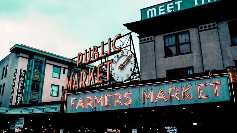 The image shows an illuminated sign reading "Public Market" and "Farmers Market" on a building facade with a clock above it, presumably at a popular market.
