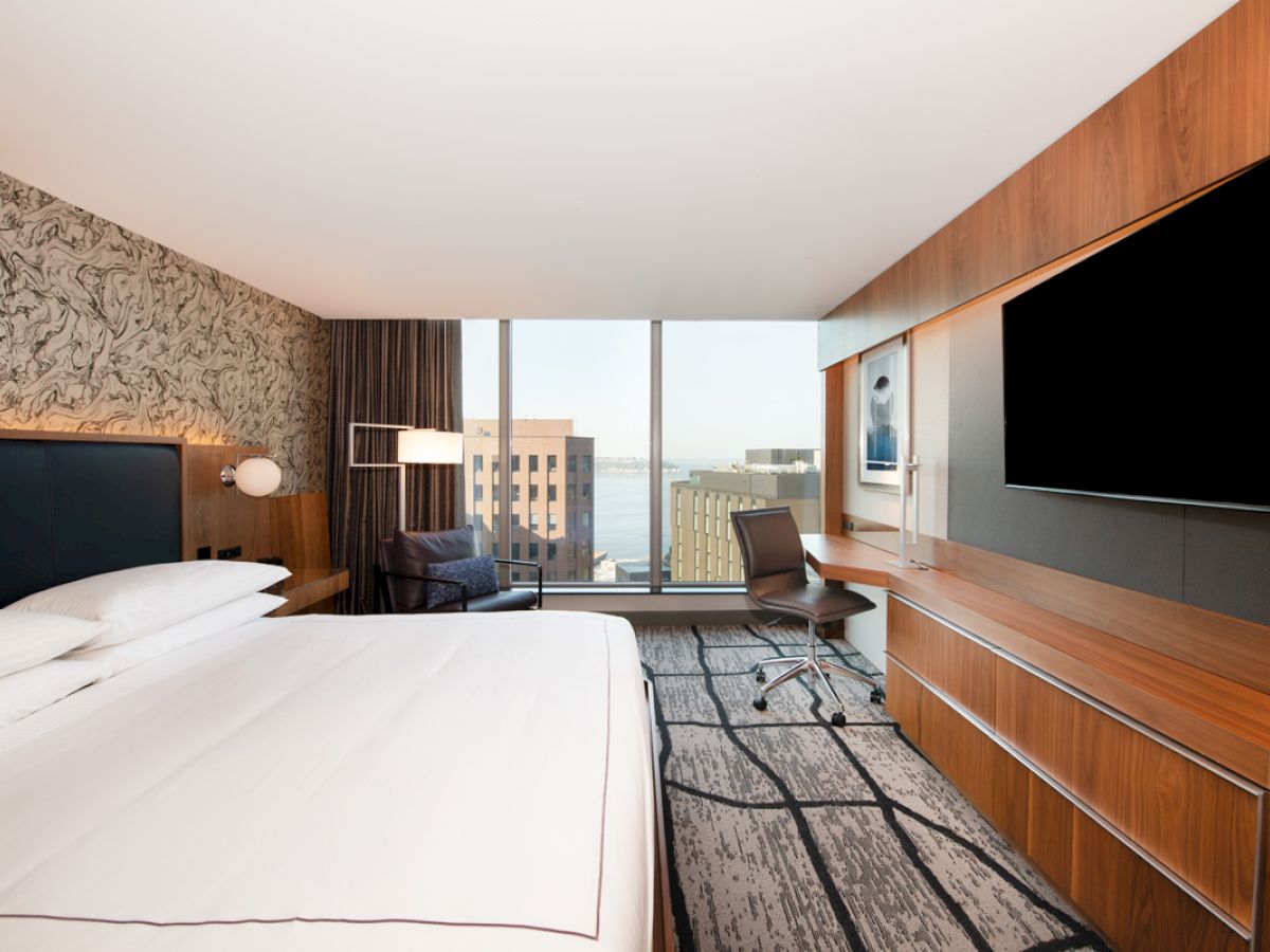 A modern hotel room with a large bed, a TV, a desk, and a chair. The window offers a cityscape view with buildings and possibly water.