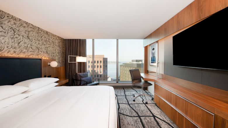A modern hotel room with a large bed, a TV, a desk, and a chair. The window offers a cityscape view with buildings and possibly water.