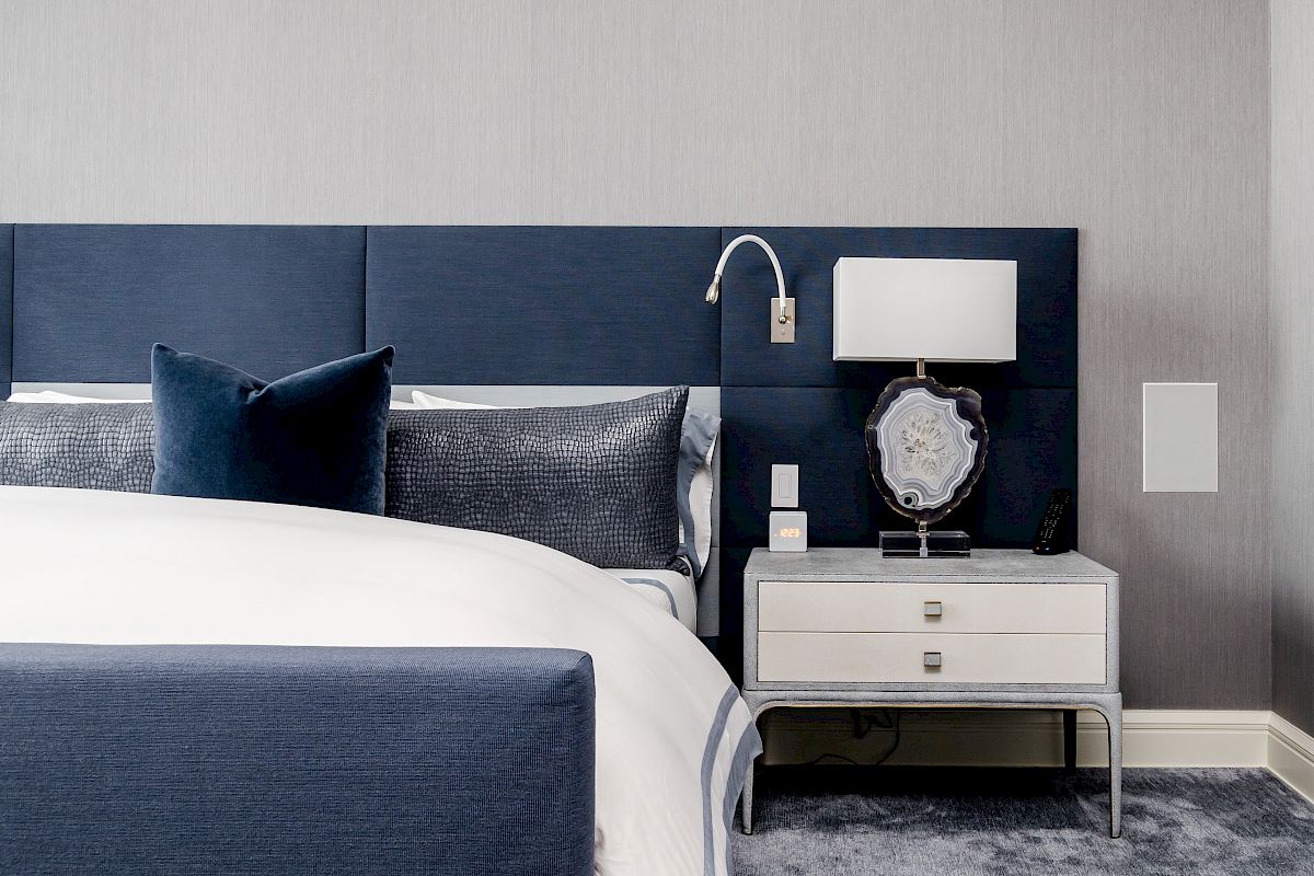 A modern bedroom features a navy blue bed with white bedding, two decorative pillows, a matching headboard, and a stylish nightstand with a lamp and decor.