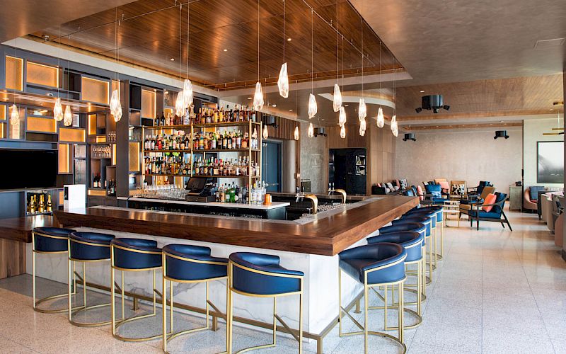 The image shows a modern bar interior with a long wooden bar counter, blue cushioned bar stools, hanging lights, and a well-stocked bar.