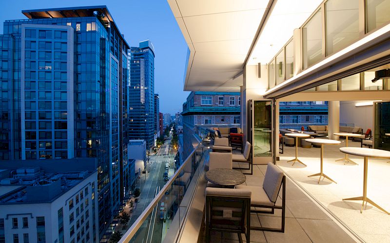 A modern balcony at twilight, featuring tables and chairs, overlooks a city street lined with tall buildings, lit by street and interior lights.