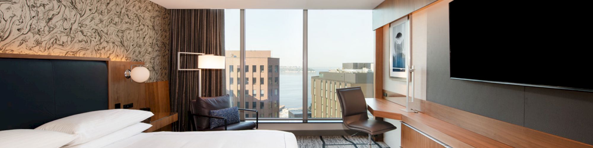 The image shows a modern hotel room with a king-sized bed, large TV, desk, and a view of buildings through floor-to-ceiling windows.