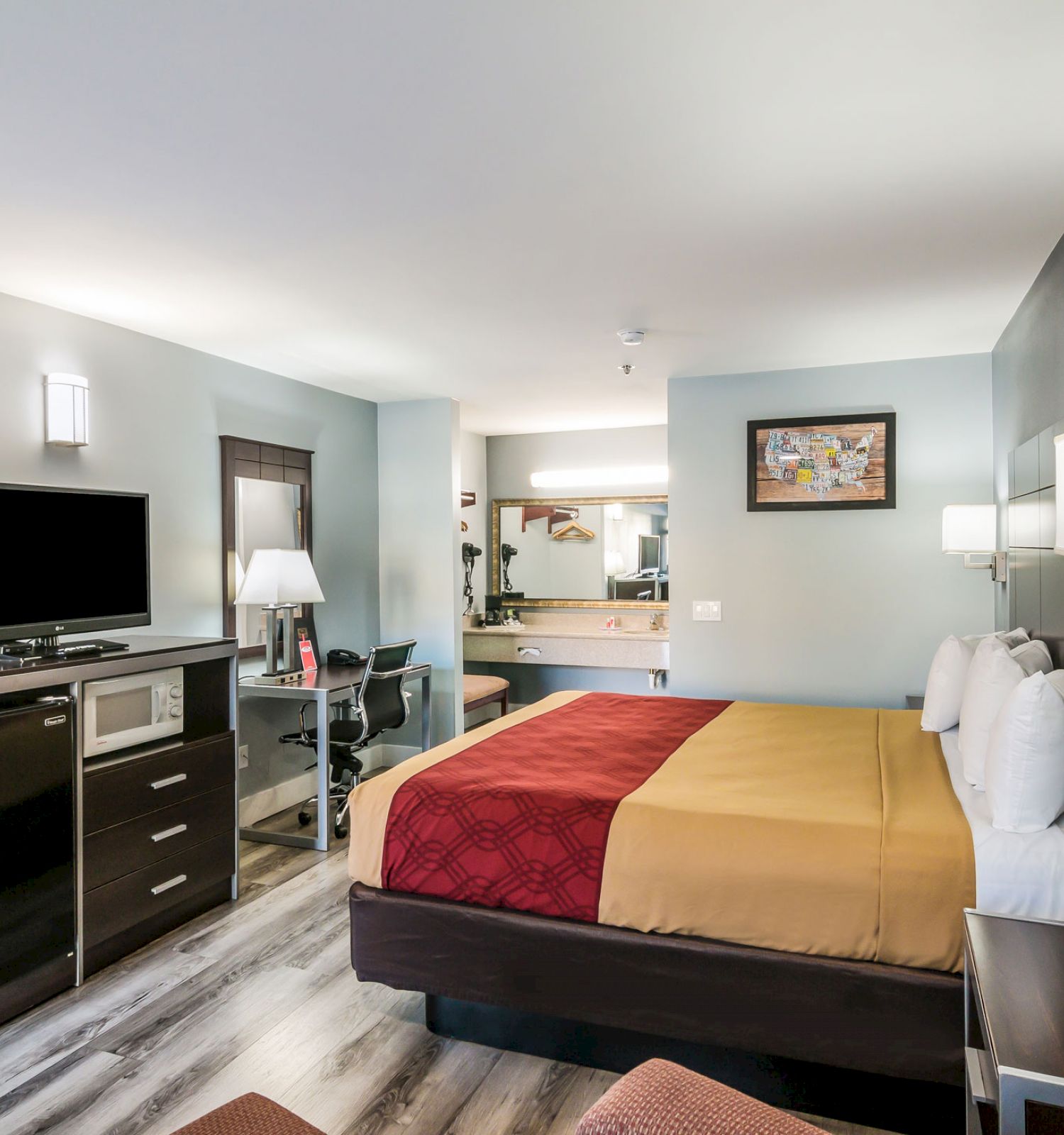 A modern hotel room with a queen-sized bed, flat-screen TV, mini-fridge, and a small workspace. The room is well-lit and neatly arranged.