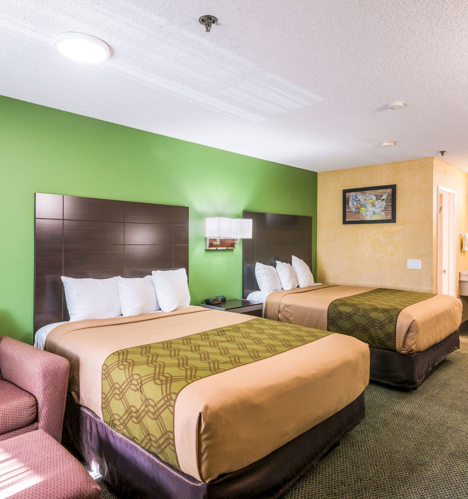 The image shows a hotel room with two double beds, a green accent wall, a lounge chair, a desk area, and a bathroom with a vanity sink area.