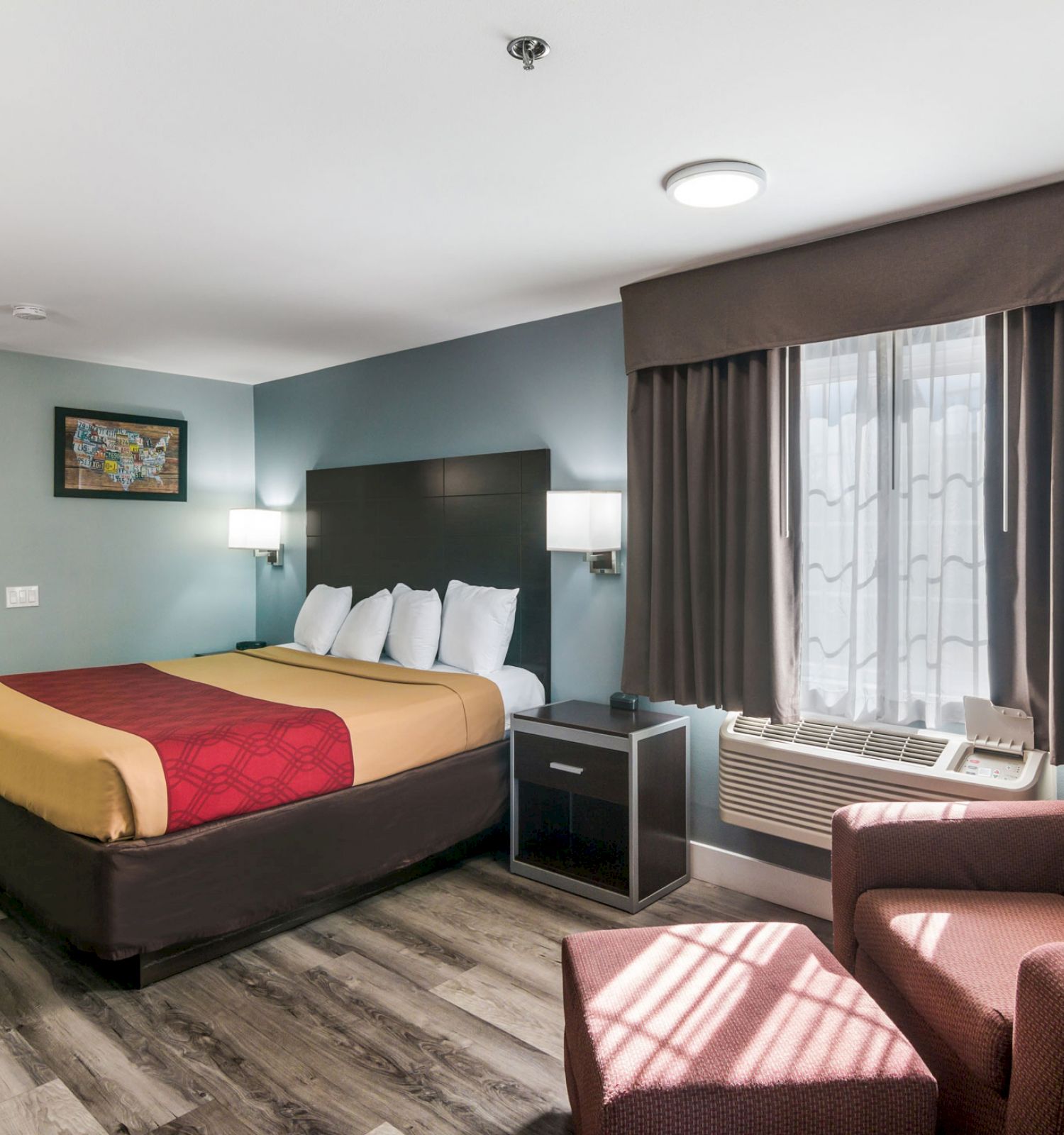 The image shows a modern hotel room with a bed, nightstand, armchair, ottoman, air conditioner, window, and wall art.