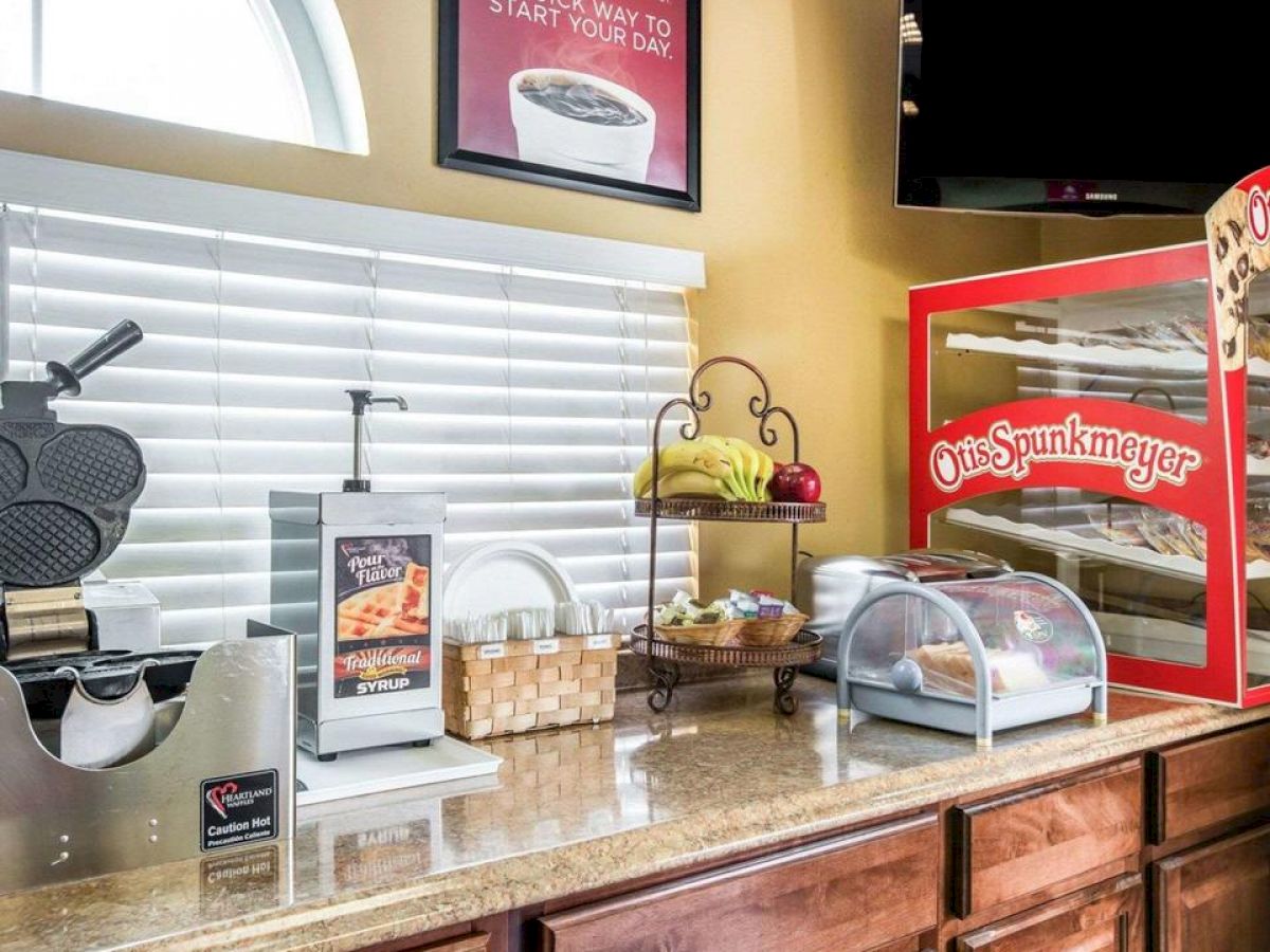 The image shows a breakfast station with a waffle maker, condiments, pastries in a display case, and wrapped muffins in a basket on a counter with a sign and window.