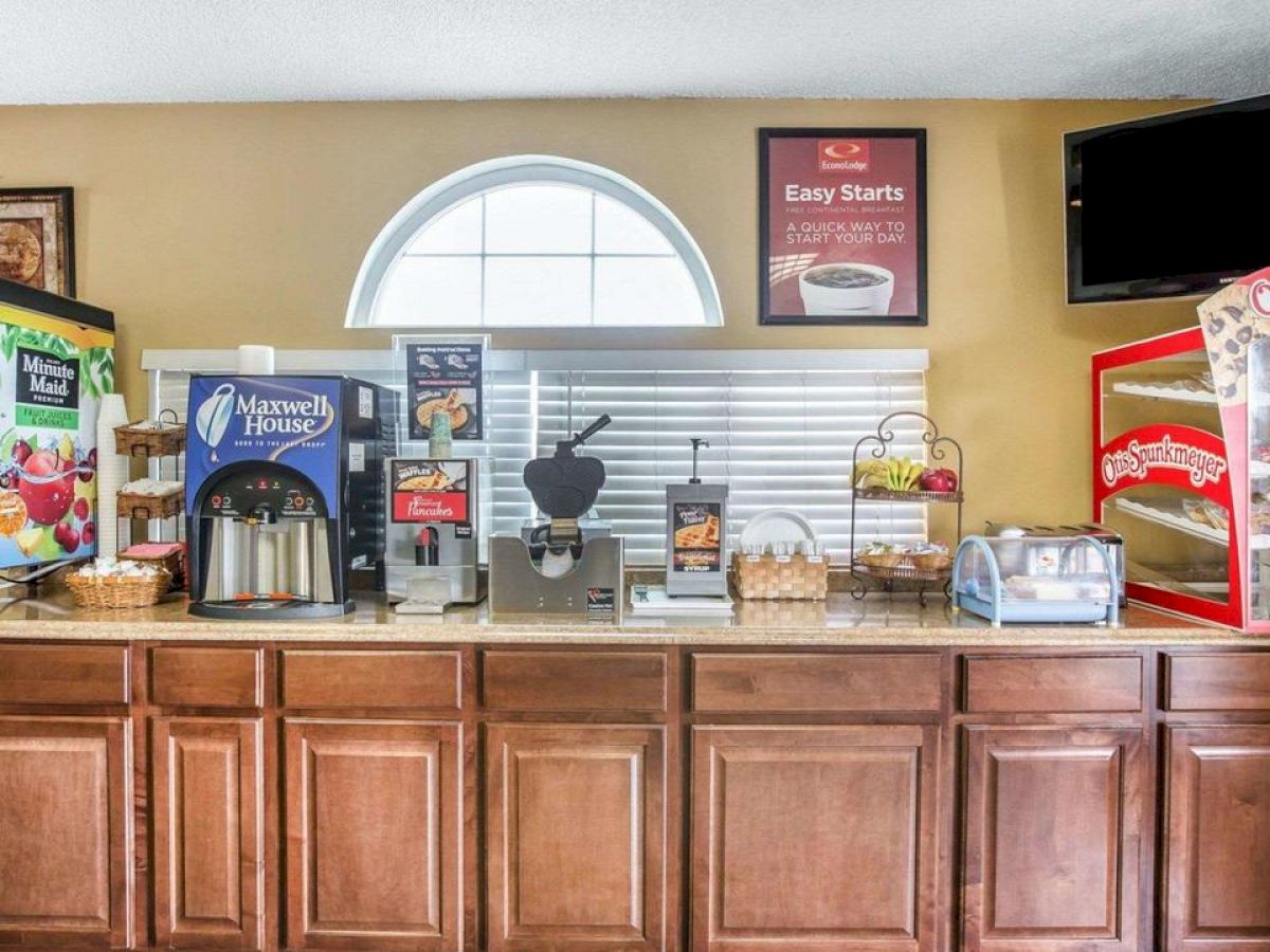 A continental breakfast setup featuring a cereal dispenser, bread toaster, Maxwell House coffee machine, and pastry trays on a counter with brown cabinets.