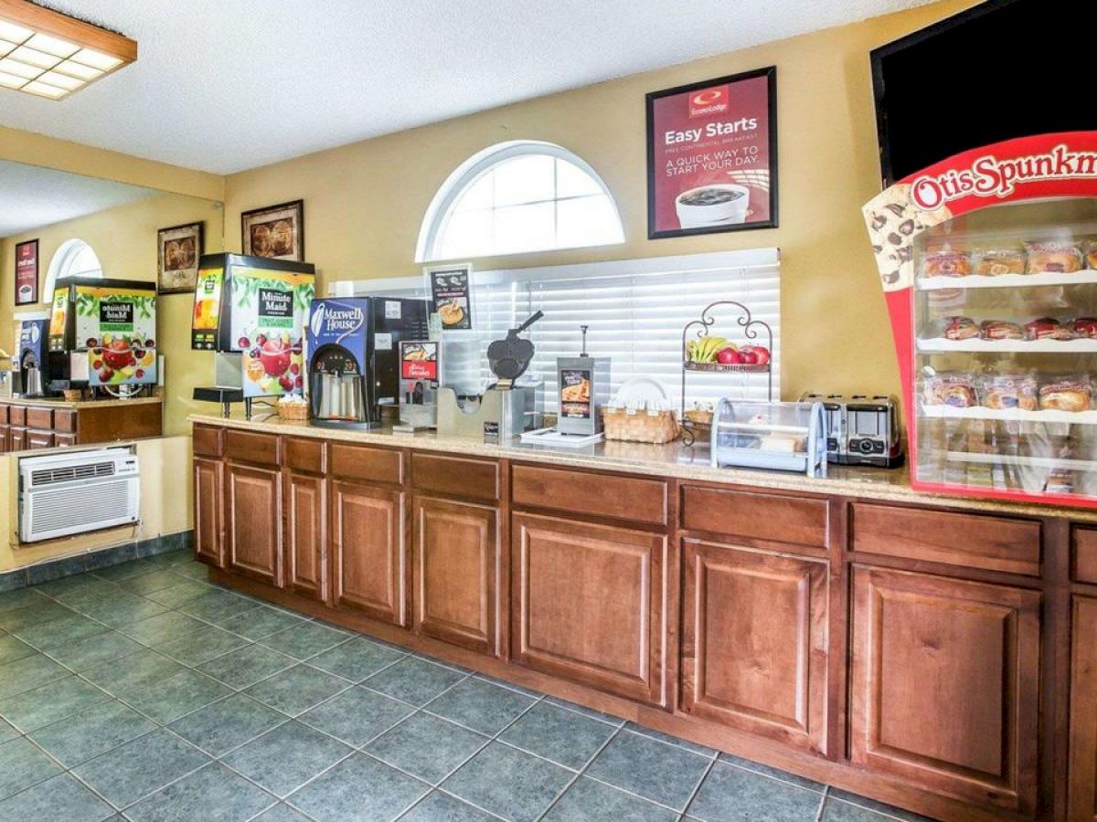 A complimentary breakfast area with various food and drink dispensers, a cereal dispenser, and a display case containing individually wrapped pastries.